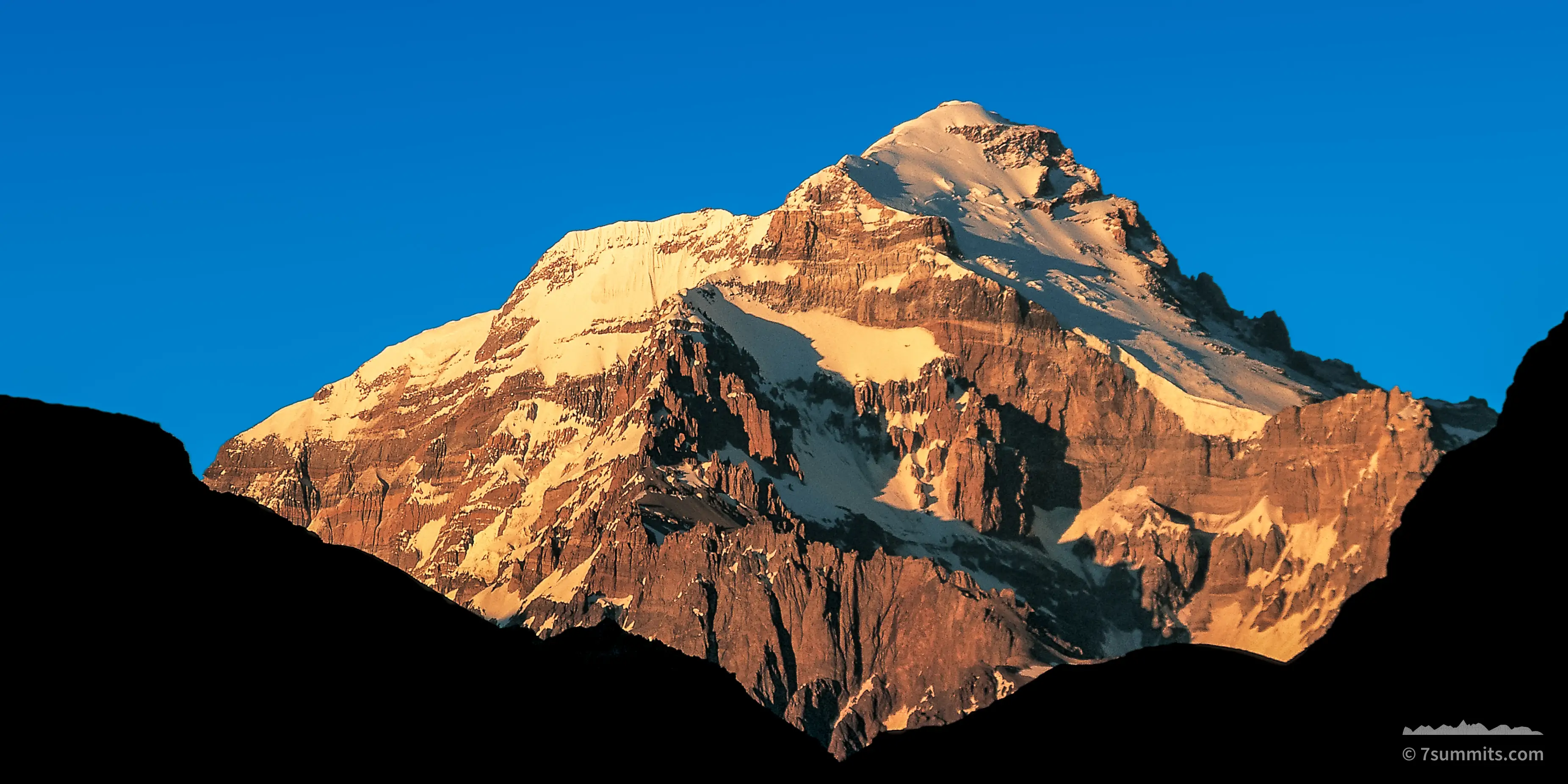 Snow and rock faces of Aconcagua lit orange by low sun, rising above black silhouetted foreground ridges under a deep blue sky.