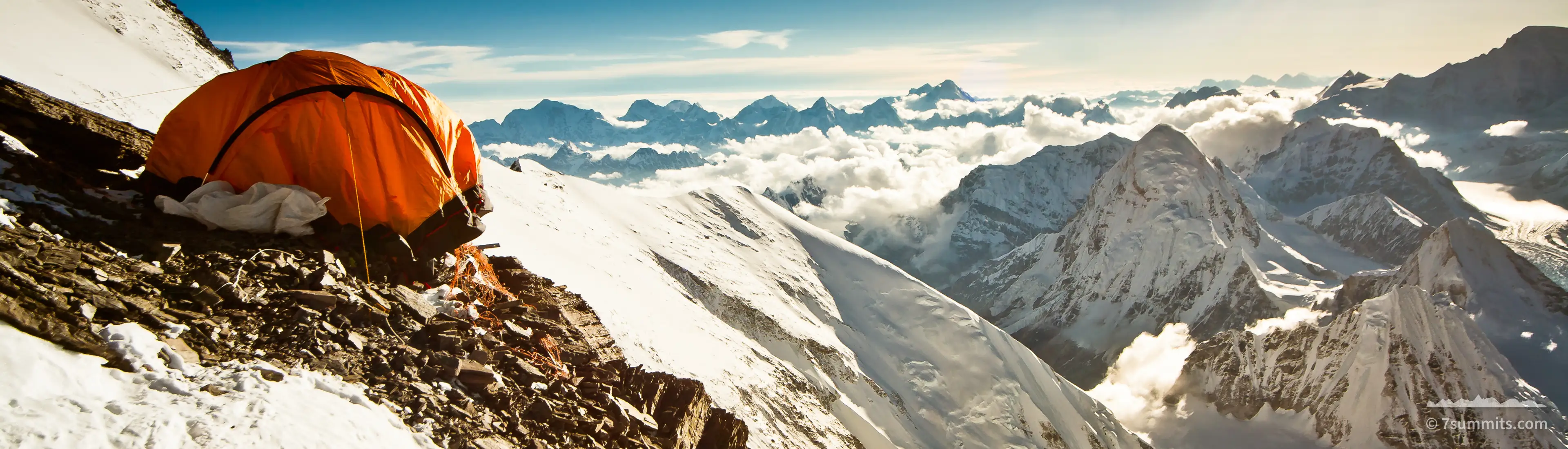 An orange single tent pitched on a rocky, snow-dusted ledge high on a mountain, with a sea of cloud and snow peaks below and low sun flaring in the upper right of a deep blue sky.