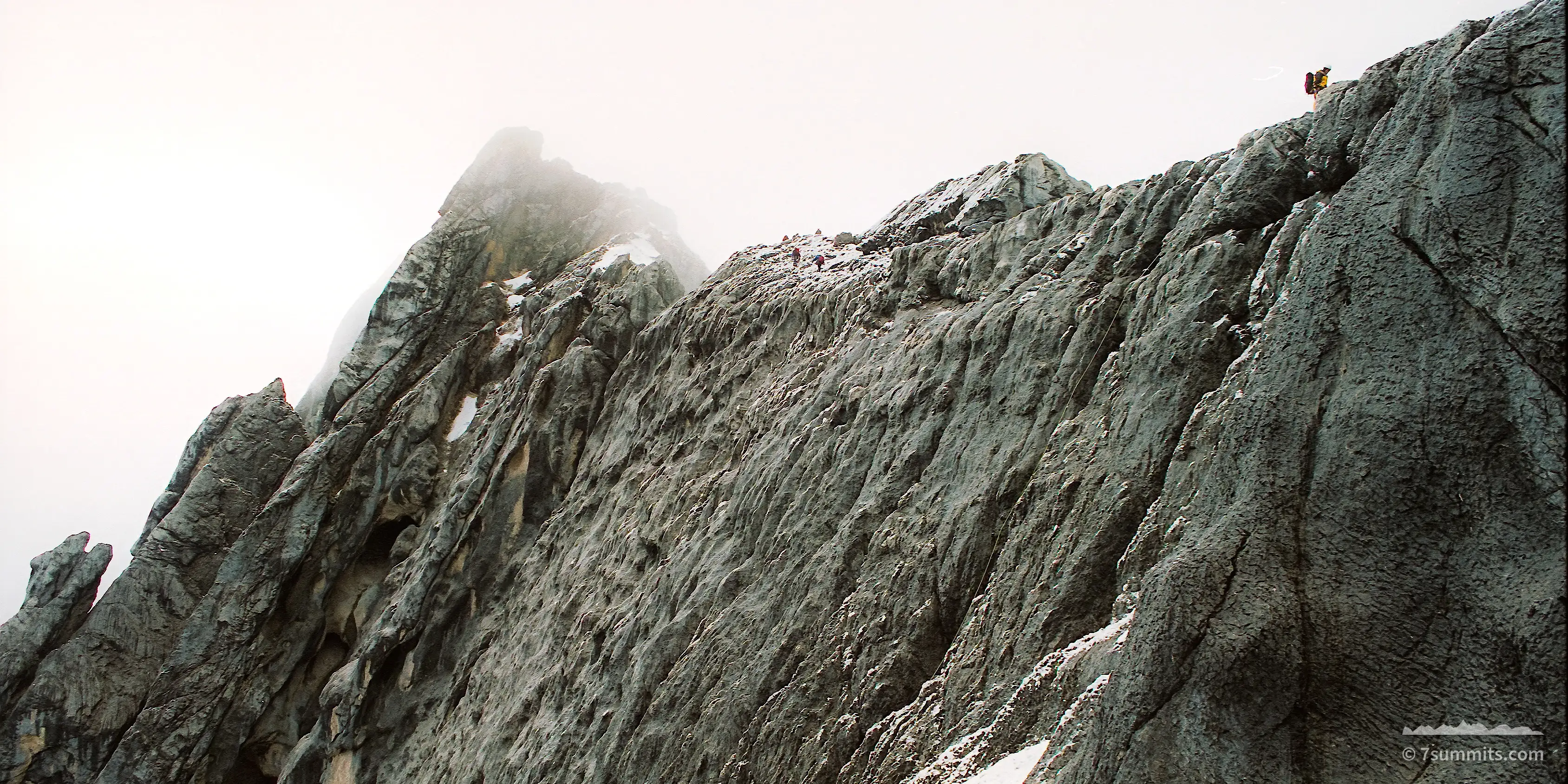 A jagged grey limestone ridge with patches of snow rises toward a mist-covered summit under a flat white sky, with a single climber in yellow barely visible on the rock at upper right.