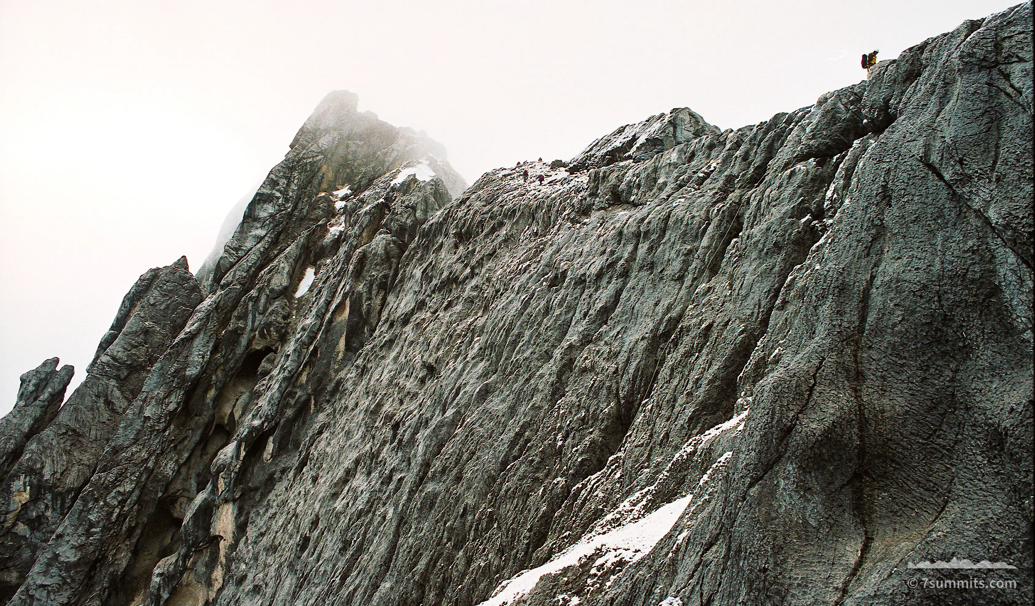 A steep grey limestone ridge with patches of snow rises into flat white cloud, with two tiny climbers visible on the crest at upper right.