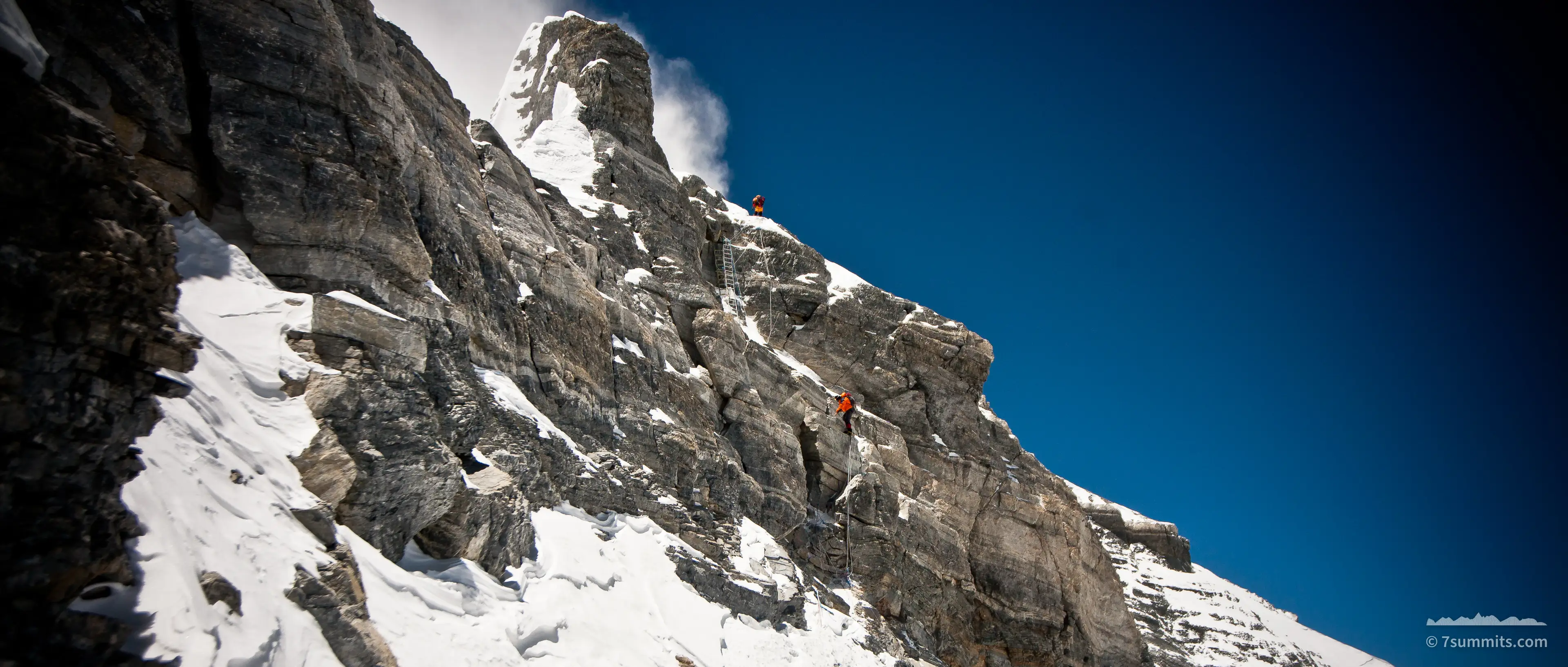 Two climbers in red suits on a snow-dusted rock buttress, one on a fixed aluminium ladder, under a deep blue sky.