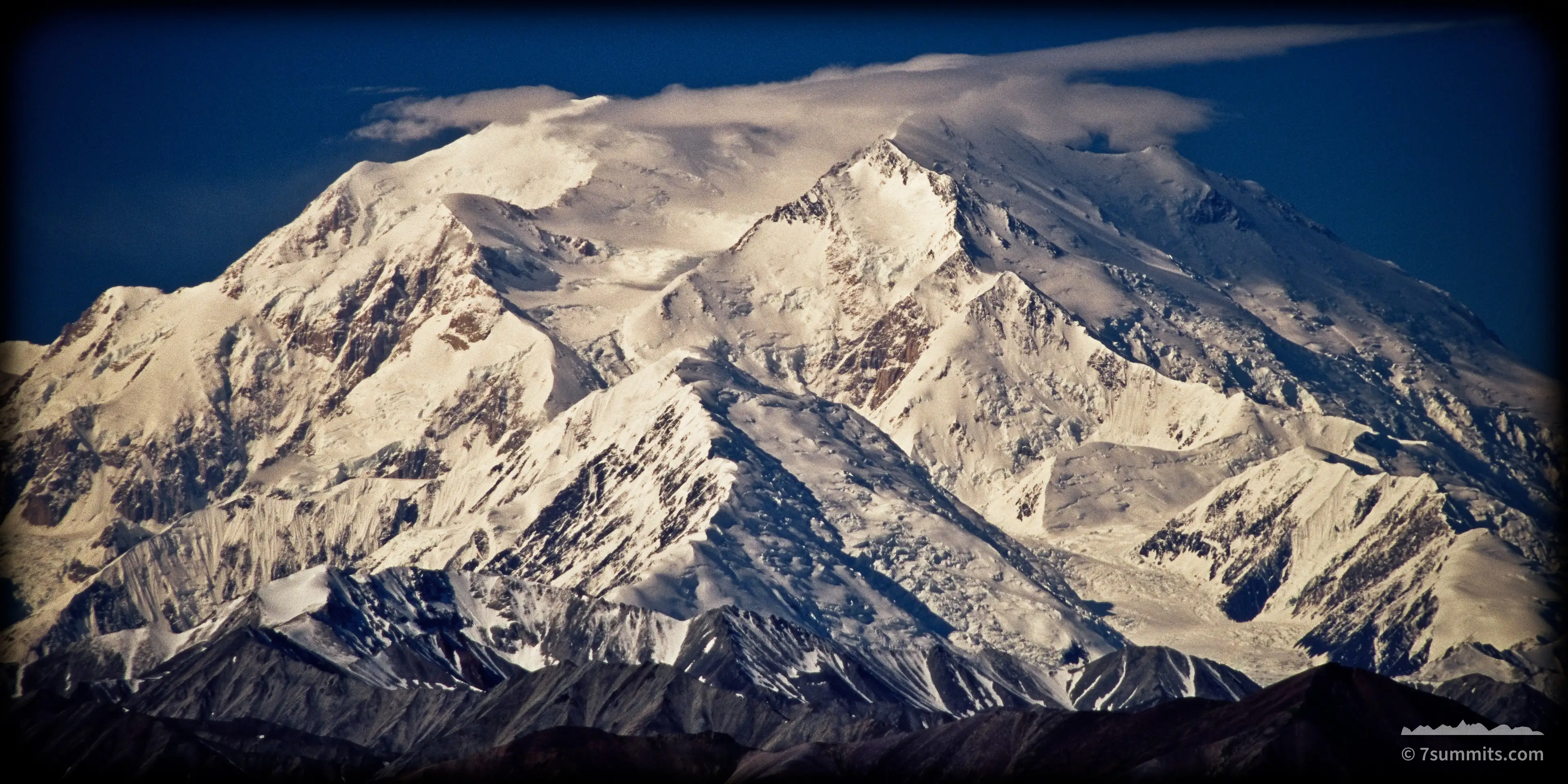 Telephoto view of Denali's snow-covered summit and buttresses under a thin lenticular cloud, with dark unlit ridges across the foreground and deep blue sky above.
