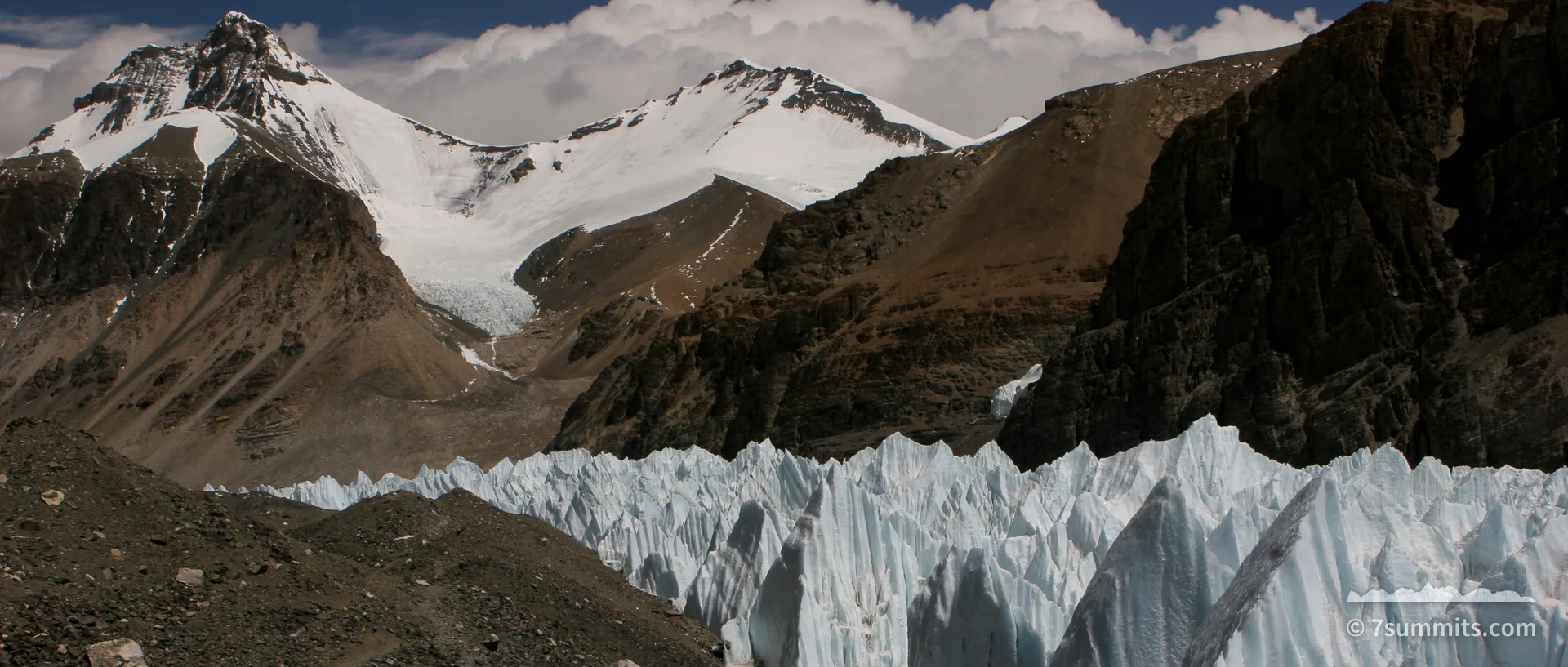 Blue ice pinnacles of a glacier rise beside a moraine ridge, with snow-covered peaks and dark rock walls behind under a partly clouded sky.