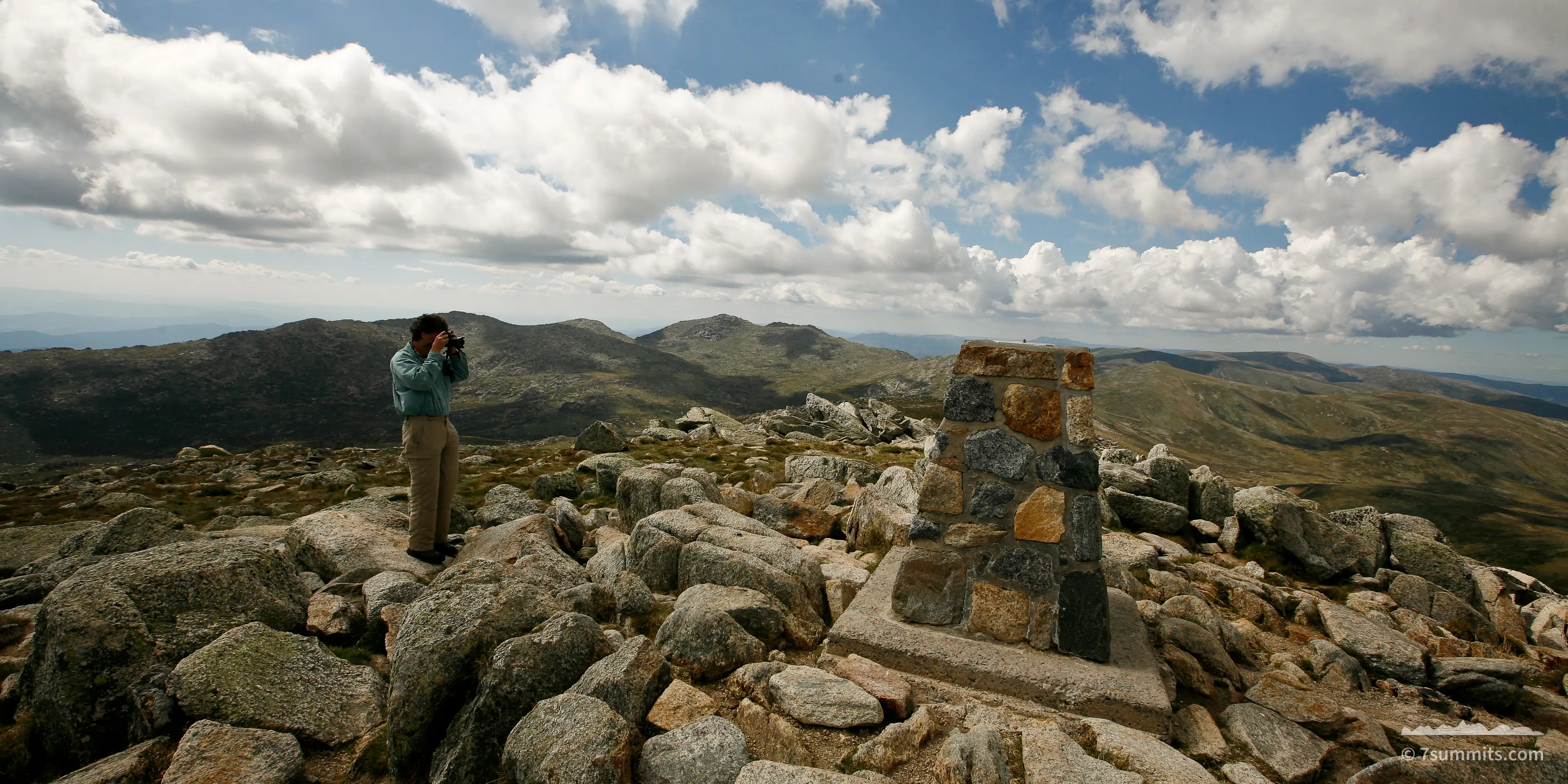 A person in a green shirt stands among granite boulders on a broad rocky summit, taking a photograph, beside a low stone cairn marker, with rounded ridges and cumulus cloud beyond.