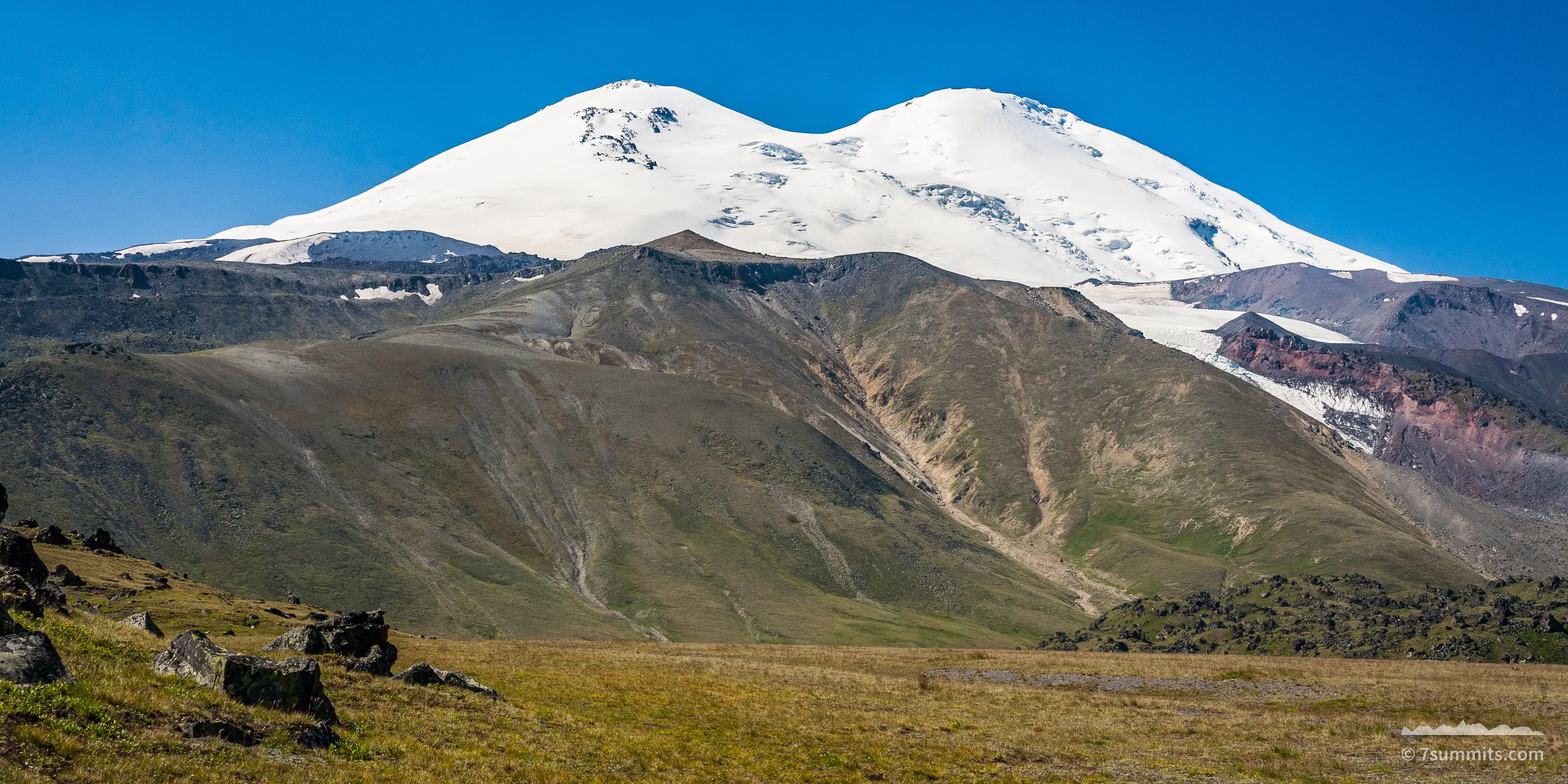 The twin snow-covered summits of Mount Elbrus rise beyond a long brown ridge and grassy foreground slope under a clear blue sky.