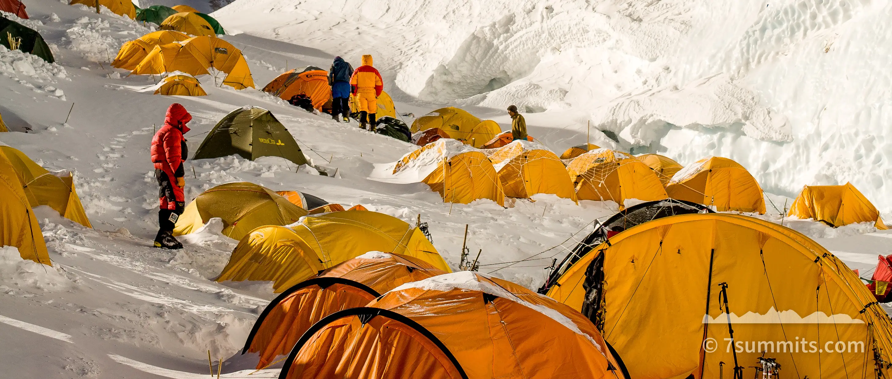 Rows of orange and yellow tents pitched on a snow slope below the wind-scoured summit pyramid of Mount Everest, with two climbers in red and yellow suits standing among them and spindrift blowing off the ridge above.