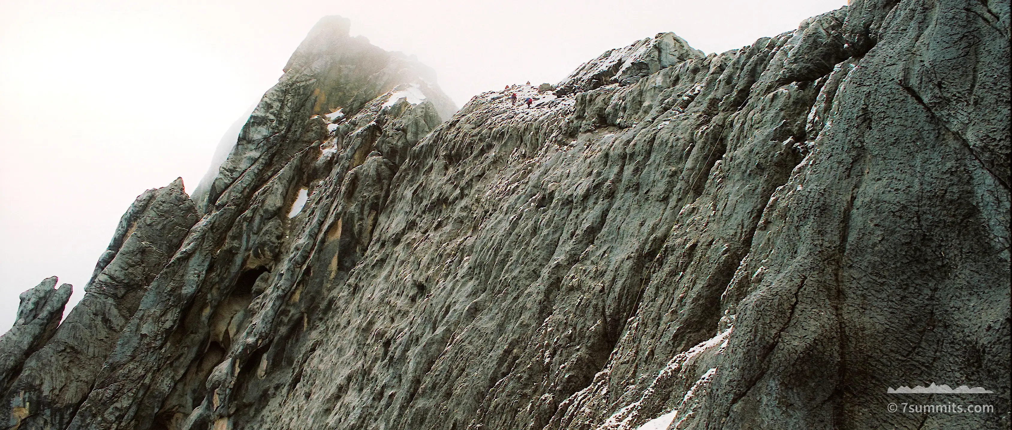 A steep grey limestone ridge with patches of old snow rises into flat white cloud, with a tiny climber visible on the crest at upper right.