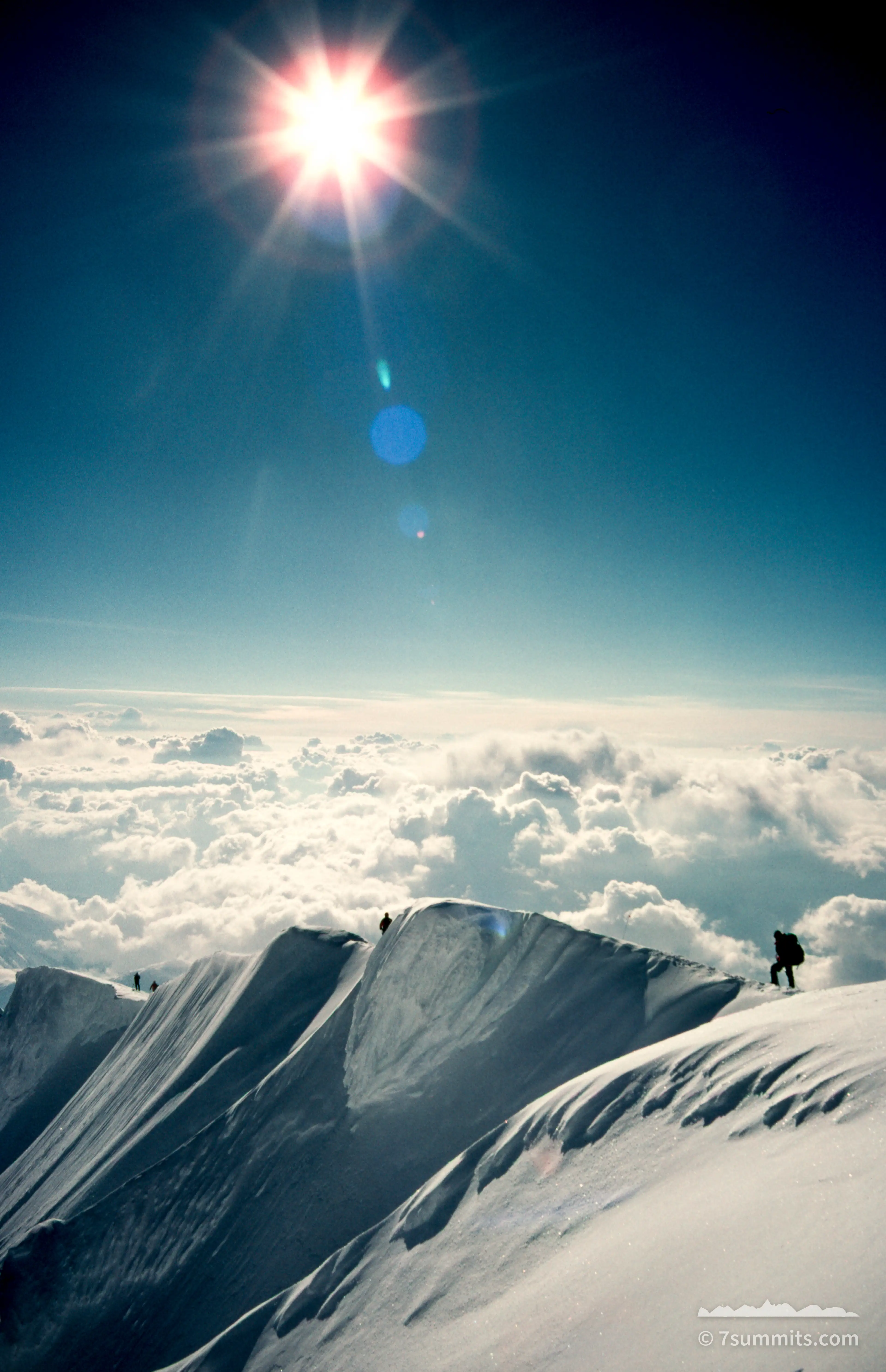 Taken from th summit of Denali, small silhouetted climbers move along a corniced snow ridge above a solid layer of cloud, with the sun flaring in a deep blue sky overhead.