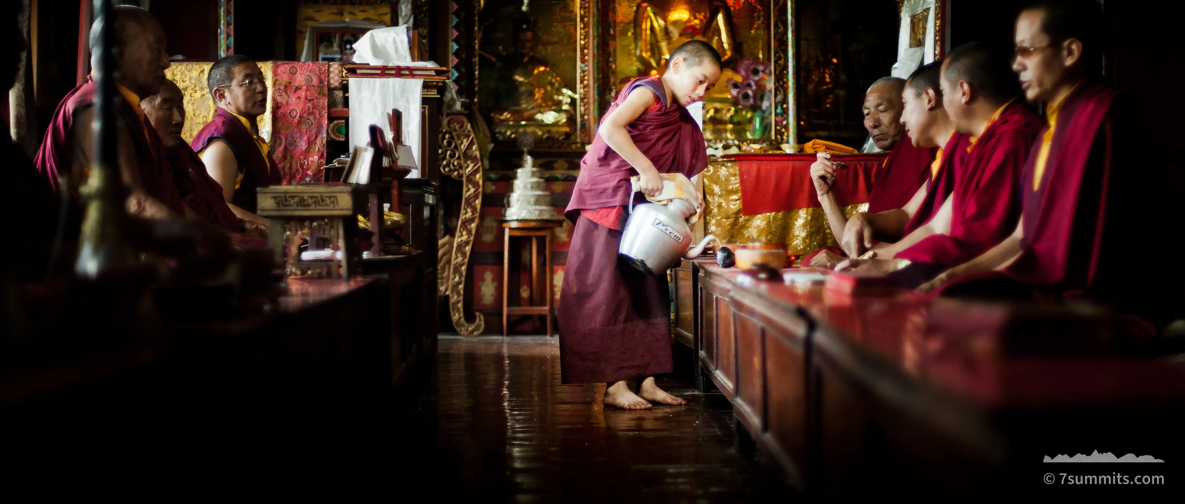Barefoot young monk in maroon robes pours tea from a metal flask for seated monks along a low table inside a dim monastery hall, with a gilded Buddha statue behind him.