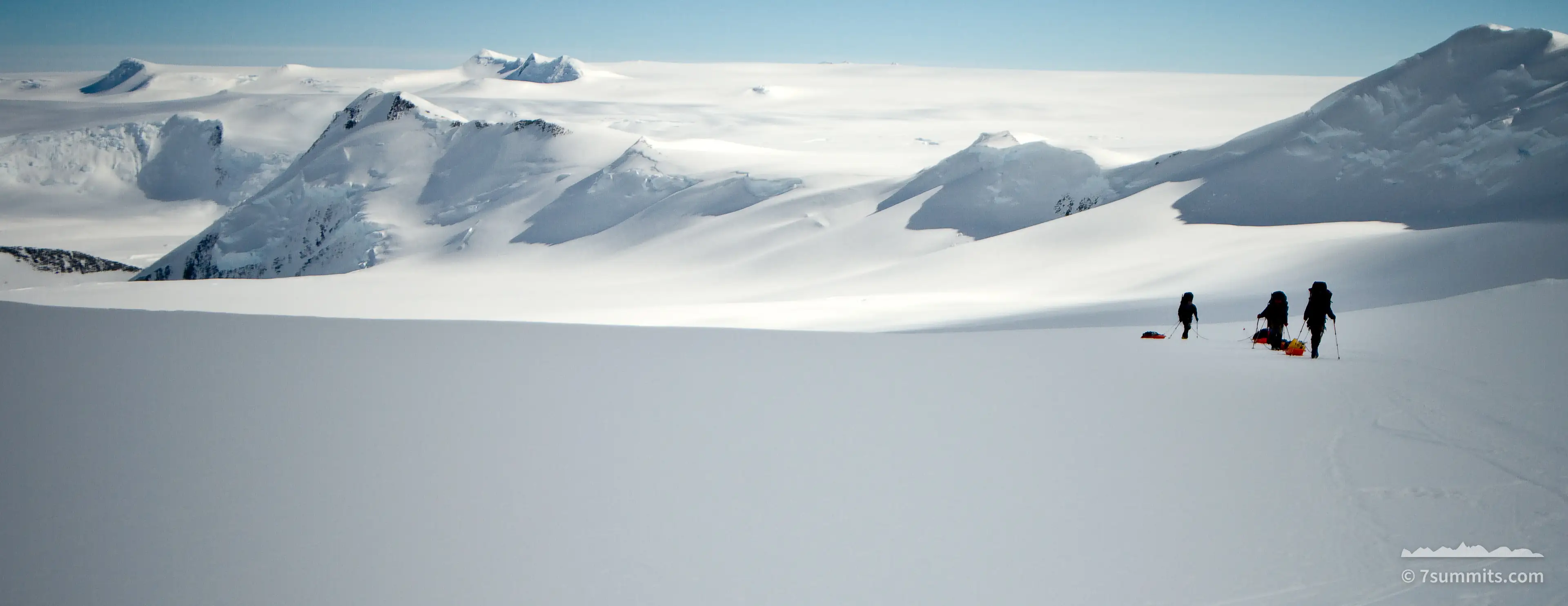 Three climbers hauling orange sleds across a broad snow plateau, with snow-covered peaks and a glacier basin behind them under a pale blue sky.