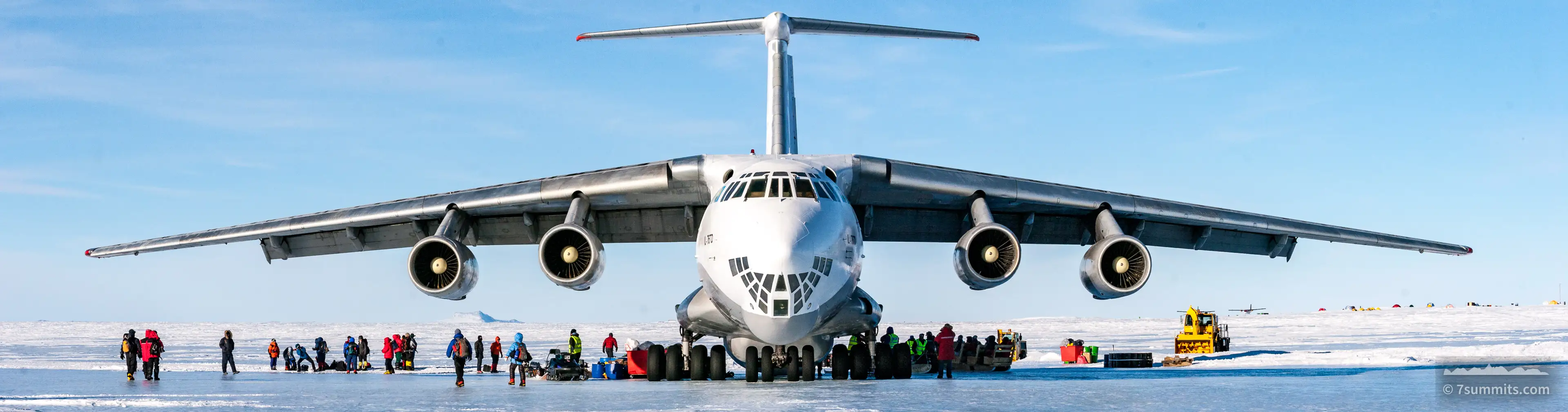 A four-engine Ilyushin cargo jet parked head-on on a blue-ice runway in Antarctica, with a line of climbers, duffel bags and vehicles unloading beneath its wings under a pale sky.